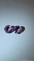 Pair of round faceted natural amethyst gemstones with pink and purple color hues, shown on a light background to illustrate clarity and luster.