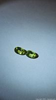 Two small oval cut green peridot gemstones placed side by side on a plain surface under lighting for clarity inspection.