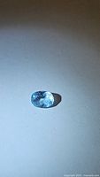 Oval cut aquamarine gemstone approximately 8x6mm with visible inclusions and light blue coloration photographed on white background.