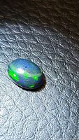 Oval shaped black opal cabochon gemstone with bright green and blue flashes on a black textured background under directional lighting.