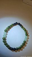 Photo of circular stretchy bracelet with alternating green jade beads, black lava stones, and green aventurine beads on a white surface taken from above.