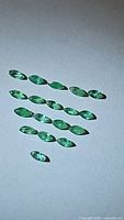 Top-down image showing 14 marquis cut emerald gemstones arranged in rows on white surface.