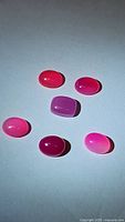 Top view of six oval cabochon cut natural hot pink chalcedony gemstones arranged on a white background with distinct shading and light reflection showing glossy surface and varying hues from hot pink to purplish pink.