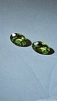 Pair of natural green peridot gemstones, oval cut, shown on neutral gray surface with light enhancing their facets and color.