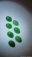 Eight green oval cabochon gemstones displayed on white background with shadows, showing different translucent green shades and polished surfaces.