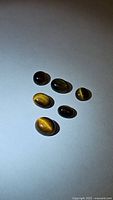 Six polished cabochon tiger's eye stones laid on a white surface showing their chatoyancy and various oval and round shapes.