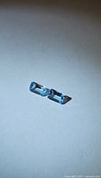 Pair of natural sky blue topaz gemstones, emerald cut, laying on a flat light surface, each showing bright blue color and clear facets.