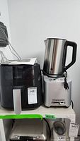 Front view showing Saltron air fryer with brand label and handle, beside stainless steel kettle and silver toaster stacked on shelf.