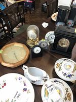 Photo showing the full lot on a dining table including the brass dome clock, wooden wall clock, mantle clock, small timers/clocks, and porcelain dishware.
