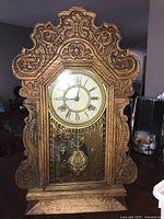 Front view of oak gingerbread clock showing detailed wood carving, clock face with Roman numerals and pendulum behind glass.