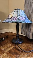 Full lamp showing stained glass shade with floral pink rose design and dark metal base on wood floor near wall corner.