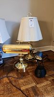 Three lamps grouped together on the floor including the brass table lamp with shade, the brass desk lamp, and the black desk lamp.