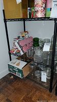 Black metal shelving unit with three wire shelves, containing various glass jars and boxes which are not included with the lot.