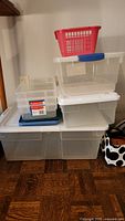 Stack of various clear plastic storage bins and one red basket on top.