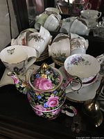 Teacups and saucers shown on cabinet with floral and butterfly designs, tea cup with matching small floral teapot in front
