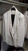 Front view of the white tuxedo jacket hanging on a hanger, showing the collar, lapel, and front pockets.