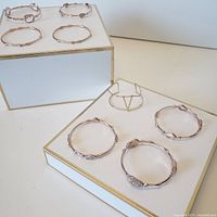 Eight gold toned bracelets displayed on white surface with gold edged stands, showing variety in design including smooth, textured and geometric styles.