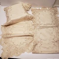 Photo showing two pieces of beige table linens with embroidered floral designs and cutwork lace scalloped edges laid flat.