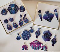 Photo showing the full set of 12 handcrafted polymer jewelry pieces arranged on white and beige board backgrounds. Includes various earrings and pins in purple and blue patterns.