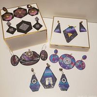Various pairs of polymer clay earrings and pendants in geometric shapes including ovals, circles, diamonds, hexagons, and triangles, in purple, black, blue, and pink tones with metallic gold and silver details.