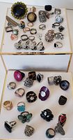 Image showing large assortment of retro and vintage rings placed on white display surfaces in two rows, featuring diverse styles and sizes.
