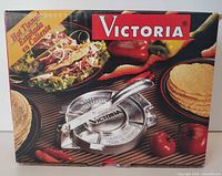 Front of the original product box showing the cast iron tortilla press image, brand name 'Victoria' and food images including tortillas and vegetables.