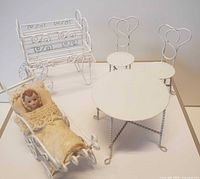 Front view of white wire dollhouse garden furniture set including bench, two chairs, round table, and pram with small doll inside.