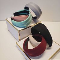 View of eight wide cloth padded headbands in various colors positioned on rectangular white platforms with gold rims.