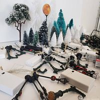 View of miniature snow-covered pine trees, an artificial snow-dusted tree, and green metal lamp posts with wiring for lights on a white surface.