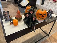 Tabletop shot showing pumpkin container, glass gourd, witch doll, scarecrow figurine, boxed decoration, candle, and ceramic haunted house