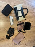 Overview showing beige tote, black case, silver wristlet, black crossbody, two blush bags, black velvet clutch on wood floor