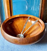 Large teakwood bowl with visible wood grain and metal salad servers inside, placed on blue carpeted surface, wooden frame and blue background.