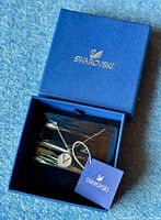 Closed-up image of Swarovski silver-tone necklace with a circular 'V' pendant encrusted with crystals inside a blue Swarovski box, with attached blue Swarovski tag.