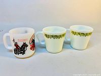 Three white milk glass souvenir mugs displayed, one featuring a colorful Royal Canadian Mounted Police design and two with green floral rims.