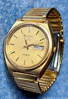 Close up angled photo of gold tone SEIKO 5 automatic men's wristwatch showing the dial with day and date display, and new gold tone stretch bracelet.