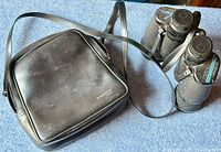 Photo of black Tasco binoculars placed next to their black carrying case with shoulder strap, showing the condition of the case and binoculars.