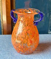 Front view of orange spatter art glass vase with cobalt blue handles against dark background.