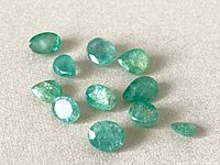 Twelve natural emerald gemstones in various mixed cuts on a white background, showing color, cut, and clarity variations.