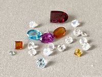 Top-down view of assortment of multicolor genuine gemstones including peridot, amethyst, moonstone, blue topaz, orange and red gems of various cuts and shapes scattered on flat surface.
