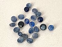 Loose blue sapphire gemstones spread out on a light surface showing various cuts, sizes, and shades of blue.