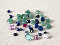Loose natural gemstones including emerald, ruby, and sapphire stones displayed on neutral background.