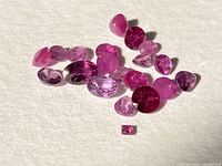 Photo showing 18 small loose genuine ruby gemstones on a white background with various pink to red colors and mixed cuts including oval, round, and teardrop.
