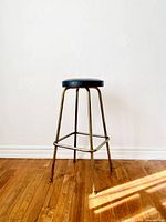 Full view of the mid-century modern high bar stool showing brass-like metal legs and black vinyl cushioned round seat on wooden floor against white wall.