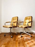 Pair of vintage Mid Century Modern swivel chairs in tan leather with wood armrests and chrome bases shown side by side on hardwood floor against white wall.