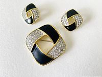 Front view of the brooch and matching earrings showing gold plating, black enamel, and clear diamante pavé in a geometric intertwined pattern.