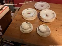 China set arranged on wooden table showing bowls, plates, cups, and saucers with floral Moss Rose pattern.