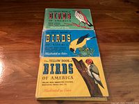 Front view of three Birds of America books stacked, showing illustrated color bird covers in green, blue, and yellow colors with titles.