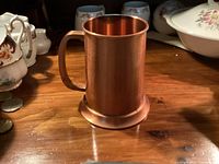 Copper beer mug on wooden surface, showing front side with handle, tarnished surface, and flat base.