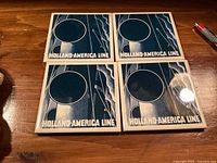 Four ceramic coasters placed on a wooden surface, each coaster has a blue and white ship design with circular indentation for glass and 'Holland-America Line' text at the bottom.