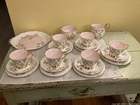 Full set showing 6 cups, saucers, small plates, cream pitcher, sugar bowl and cake plate arranged on table with white linen. The pastel pink and olive pattern with gold trim is visible.